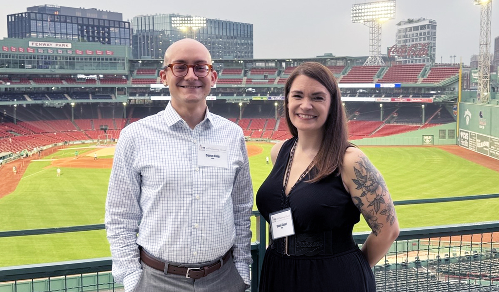 Two people smiling for a photo at Fenway Park with the baseball field and stadium seats in the background.