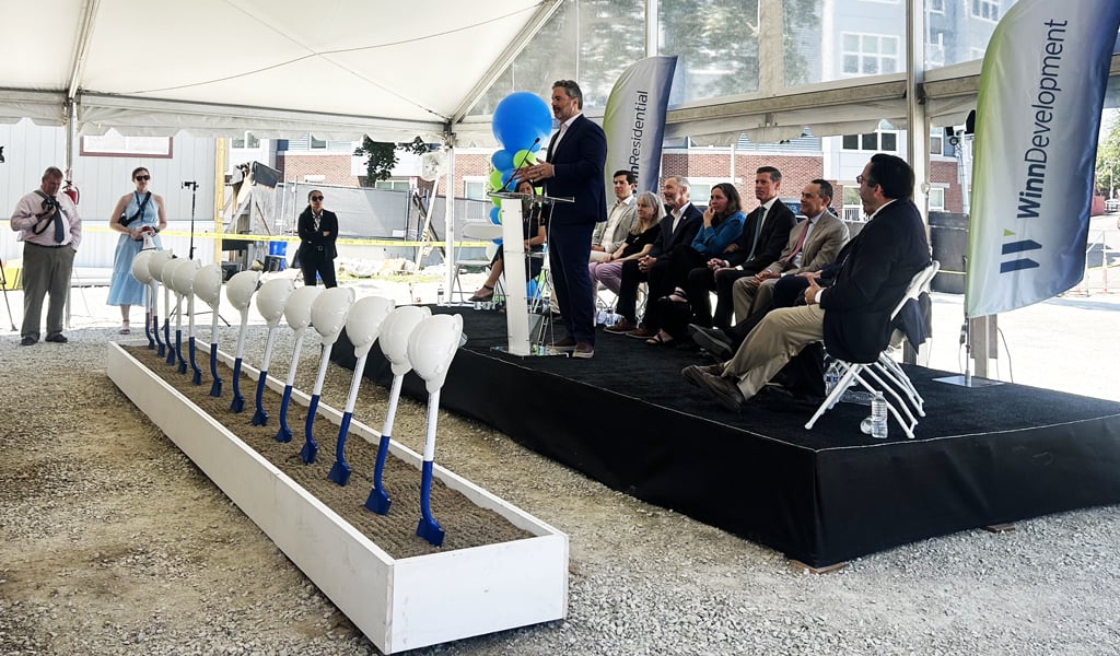 A group of individuals seated under a tent at a groundbreaking ceremony with shovels lined up in front, and a speaker at a podium.