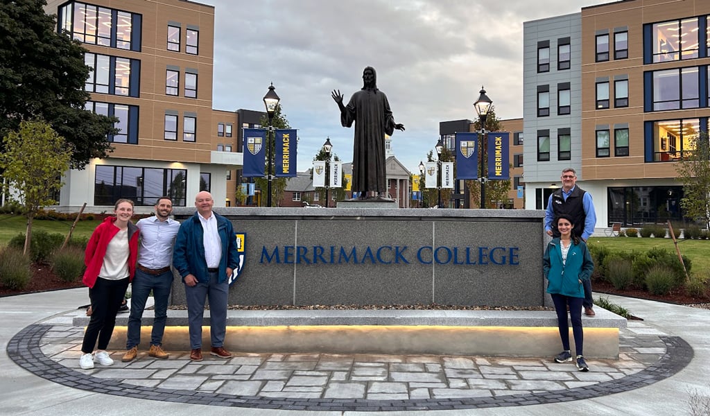 The image shows a group of five people standing in front of a sign for Merrimack College. Behind them, there is a large statue with modern campus buildings in the background.  