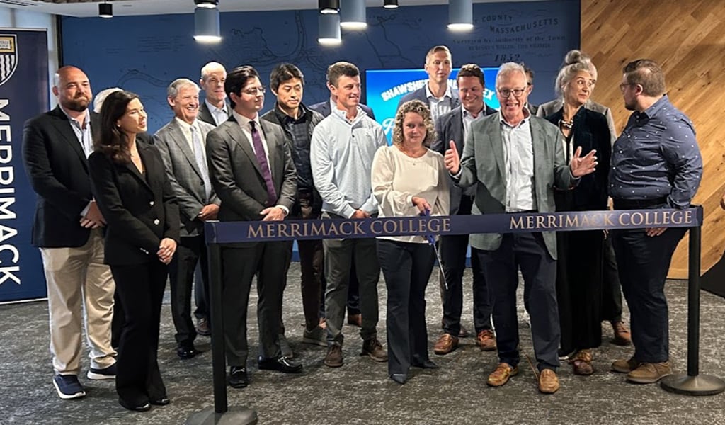 A group of people stands indoors for a photo next to a ribbon labeled "Merrimack College." They are in front of a blue backdrop with a Merrimack College logo.  