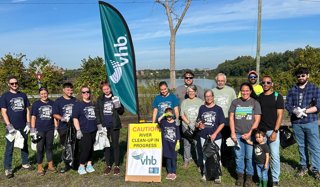 Group of volunteers with VHB banners participating in a river cleanup, holding trash bags and tools, standing outdoors near a water body on a sunny day.