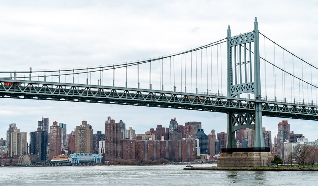 Suspension bridge over water, with the New York City skyline in the background under an overcast sky.