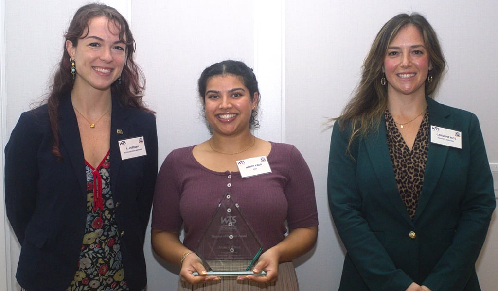 Three people stand together and smile at the camera while the woman in the center holds a framed award.