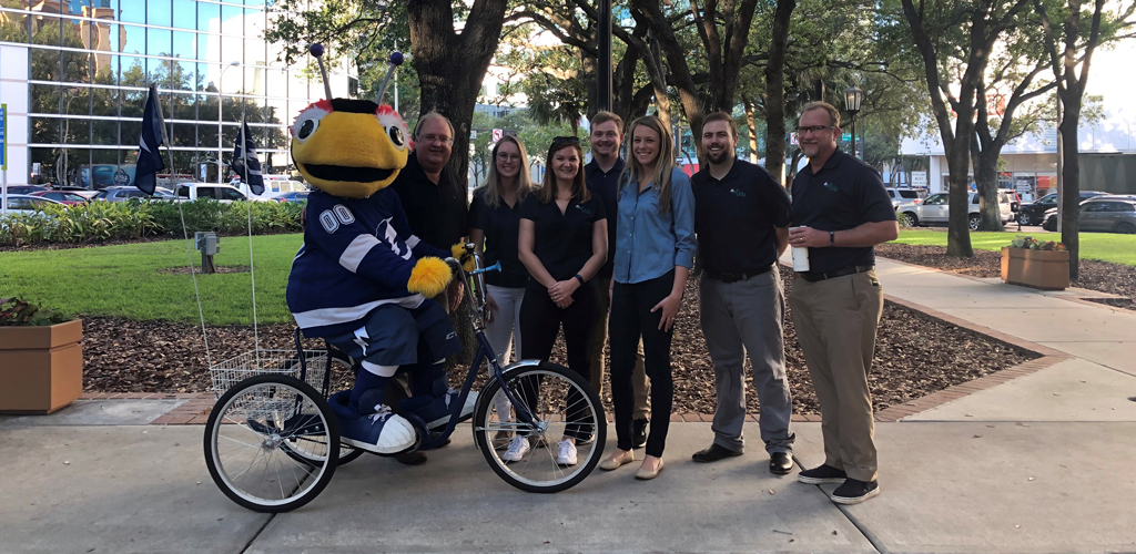 Neale and a group of co-workers stand outside an office building by the Tampa Bay Lightning ThunderBug mascot that is riding a bicycle