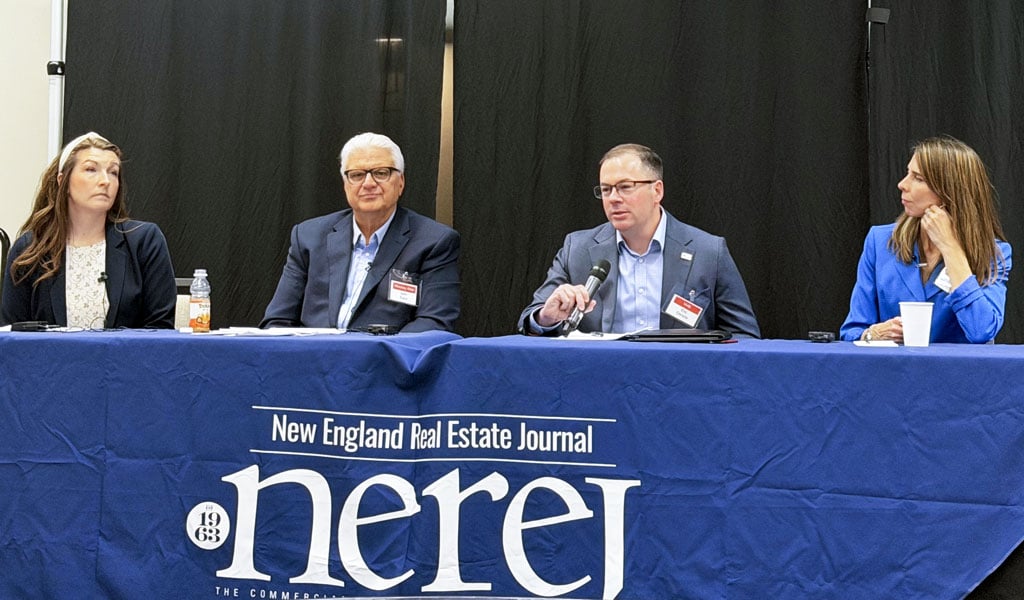 A panel of four individuals with microphones is seated at a table covered with a New England Real Estate Journal tablecloth. 