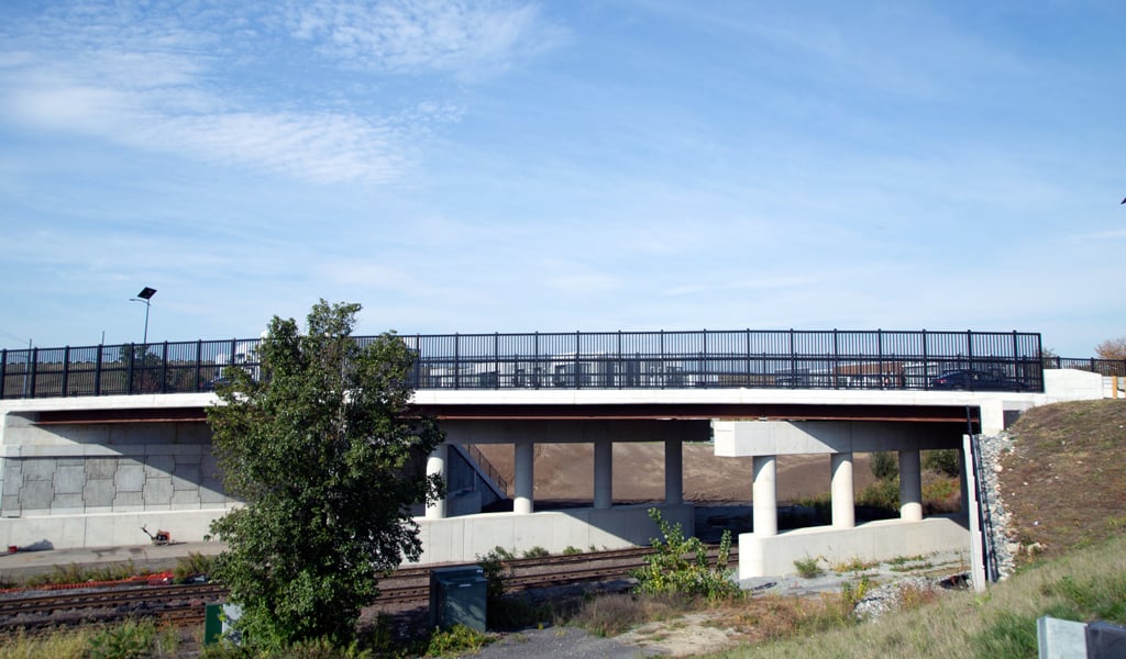 A modern bridge with a black metal railing crosses over train tracks. A small tree and some greenery are visible near the base of the bridge.