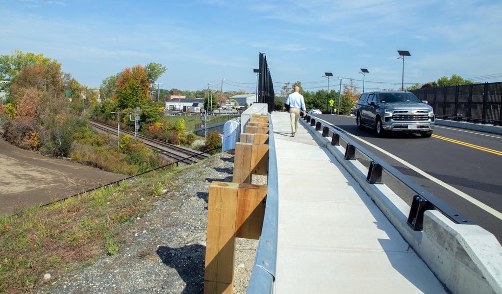 A person walks on a sidewalk next to a road with a passing SUV. The scene includes a railroad track and a variety of trees with autumn foliage. The sky is clear and blue.