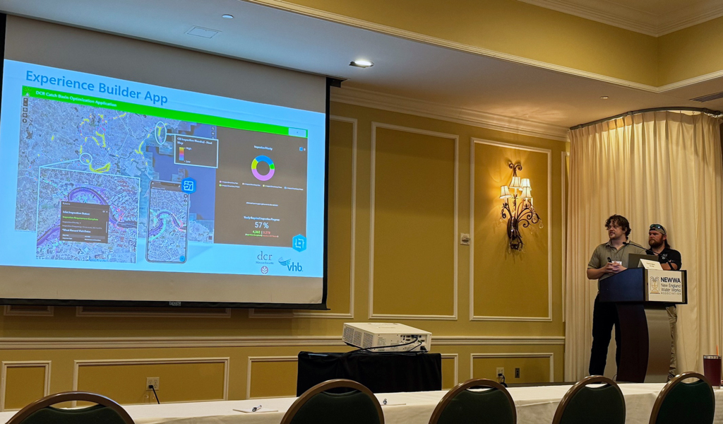 Two individuals presenting at a conference, standing beside a screen displaying the "Experience Builder App" with graphical data and maps.  