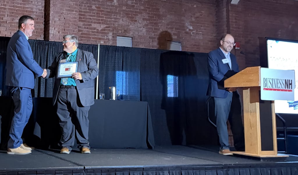 Two people are shaking hands on stage at a Business NH Magazine event. One is receiving a plaque. Another person stands at a podium with a microphone.