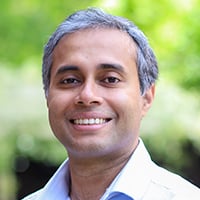 A person wearing a light button-down shirt is smiling outside with trees in the background.