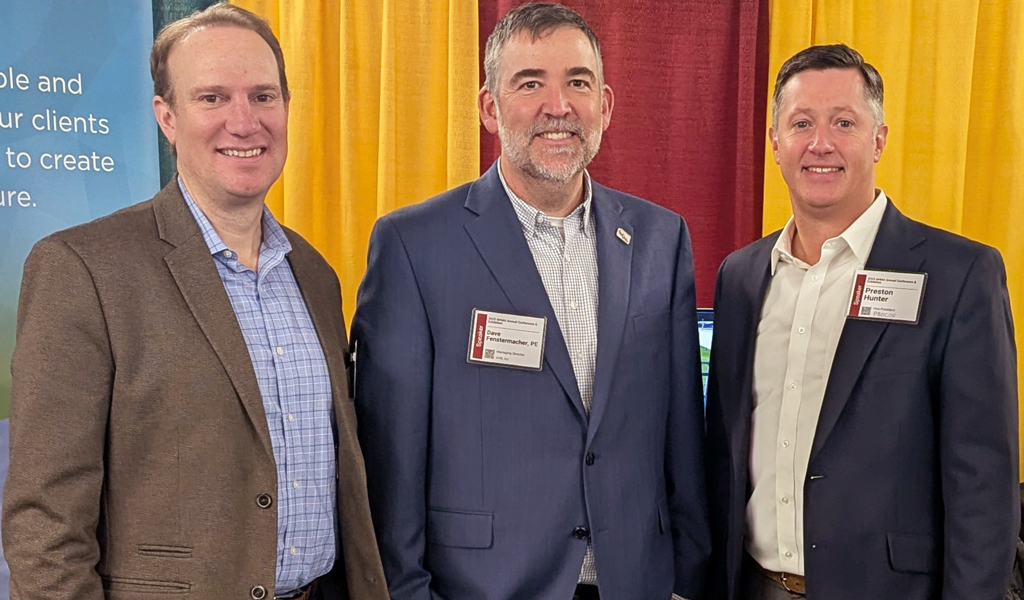 Three individuals standing in front of a banner for VHB, Inc. at a trade show booth. The banner text promotes sustainability and partnership.