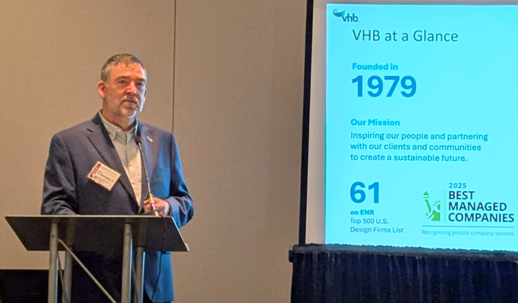 A person stands beside a projector screen displaying a presentation titled "VHB at a Glance," highlighting the company's founding in 1979 and various accomplishments, in a conference room setting.