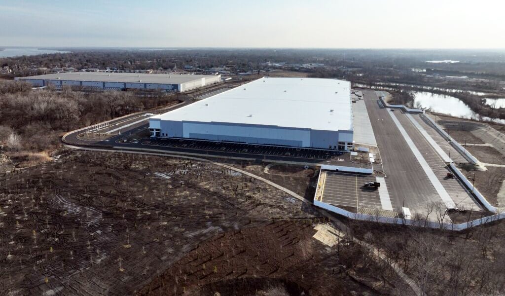 Aerial view of a large white logistics warehouse with parking areas, access roads, and surrounding wetlands.
