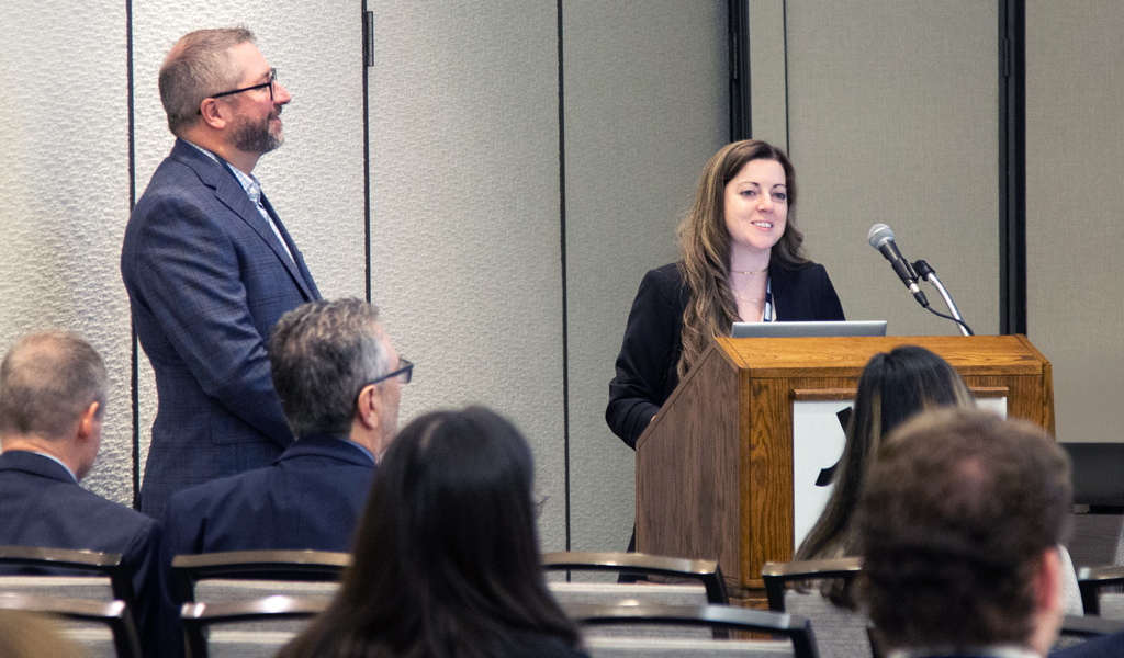 A person stands at a podium with a microphone, addressing an audience in a conference room. Another person stands beside them. 