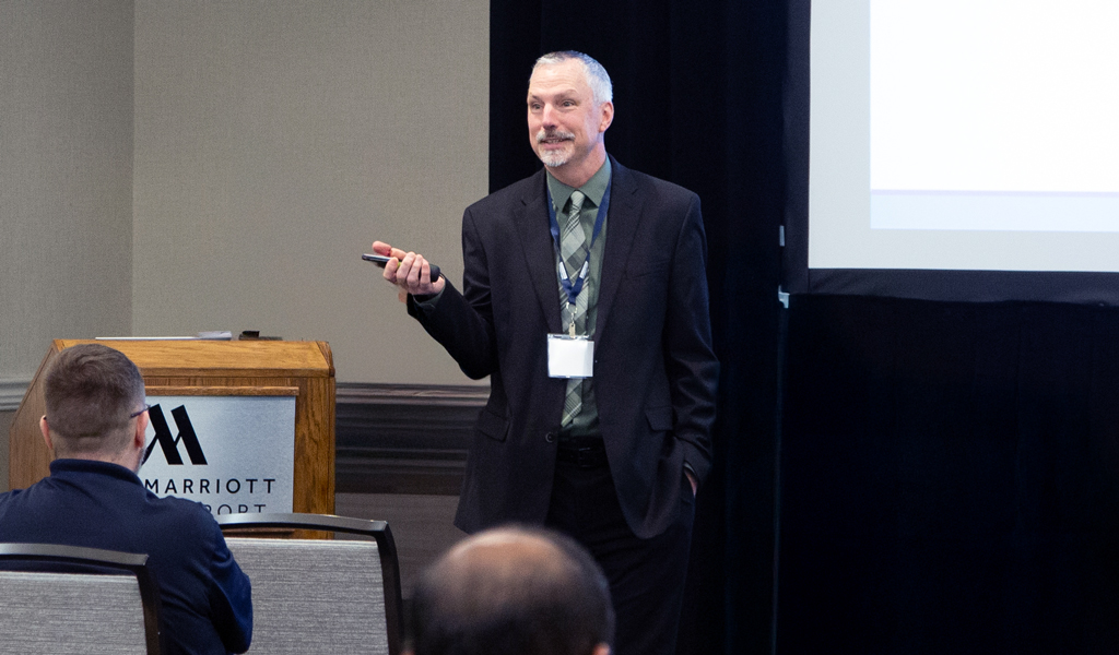 A person stands at the front of a conference room with a Marriott podium, gesturing while presenting a slideshow.