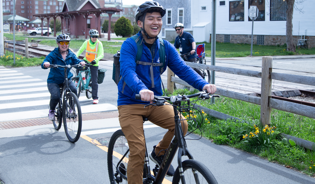 Cyclists ride along a pathway near a stop sign, with one person leading. Behind them, others follow on bikes.  