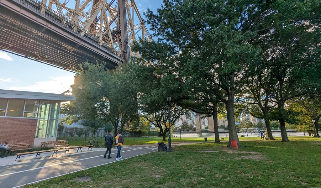 Park scene beneath the Queensboro Bridge with people walking along a path, surrounded by trees and river and city views.