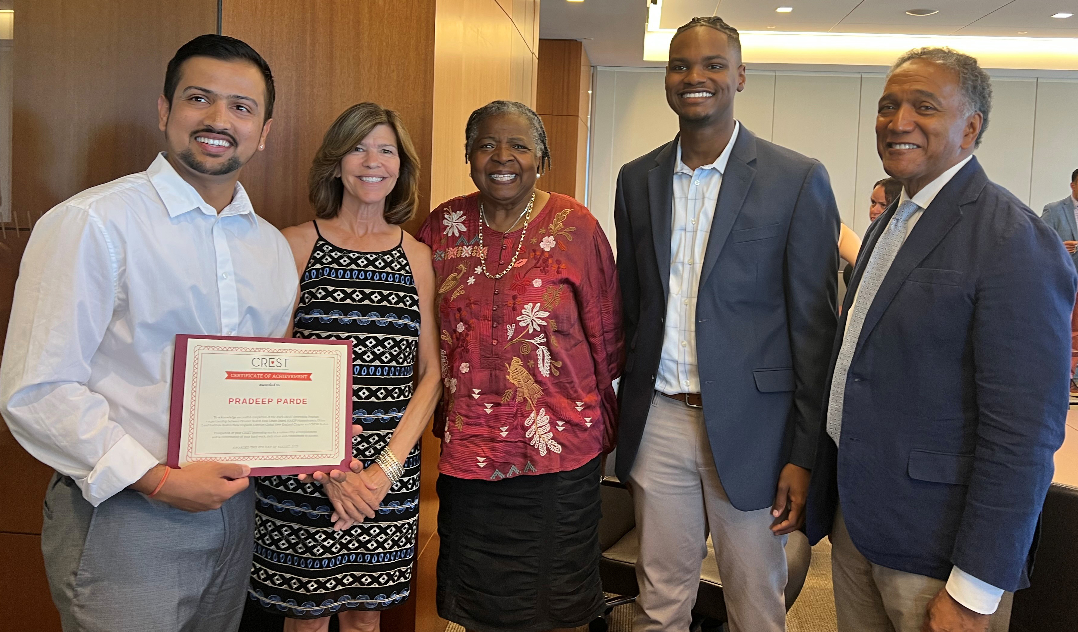 A group of people in business attire stands in an office. One person holds a framed certificate, and the group poses together.