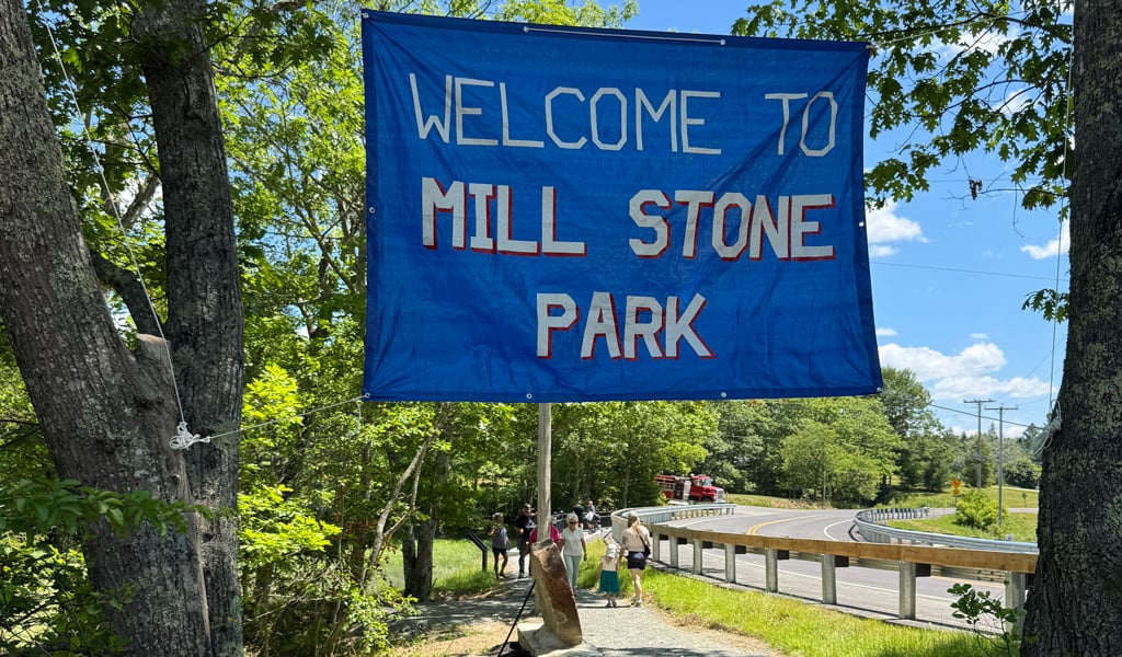 Welcome to Mill Stone Park” banner hanging over a shaded gravel path near the road and trees.
