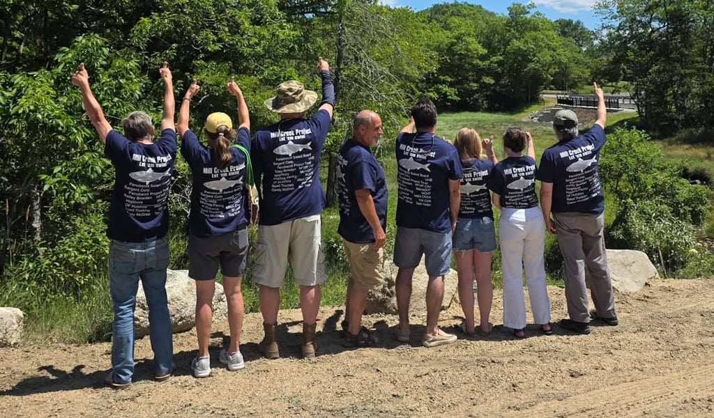 Group in matching shirts raising their arms on a sunny dirt path by the water and trees.