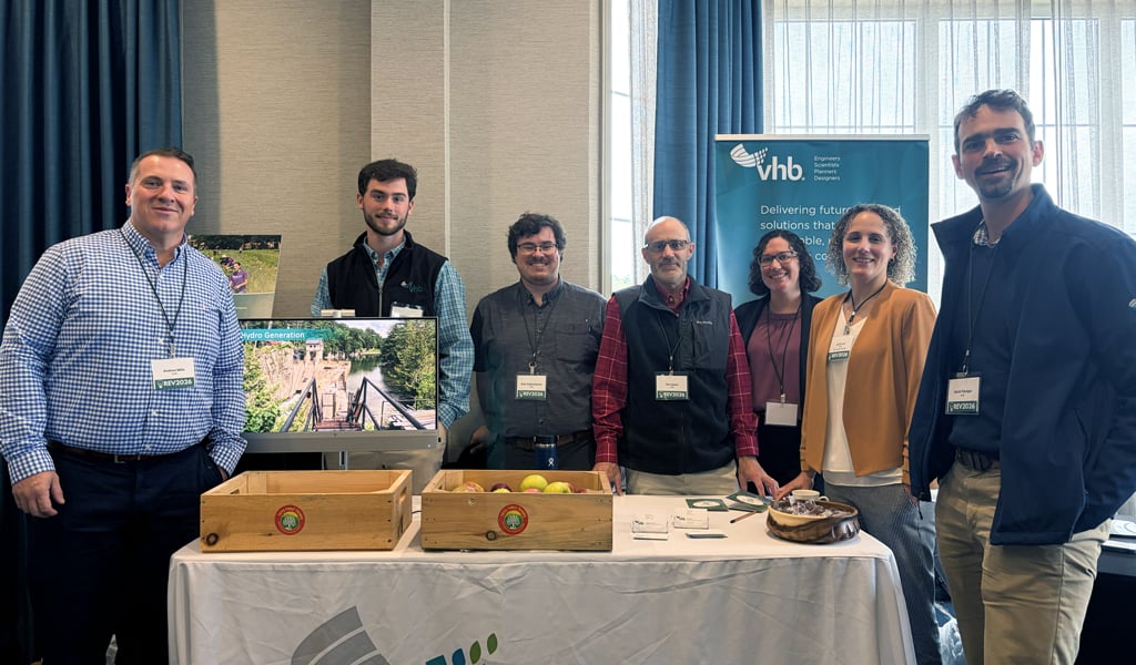 A group of individuals stands behind a table with a VHB banner. The table displays boxes of apples and informational materials. A screen in the background shows a scenic image of a trail.