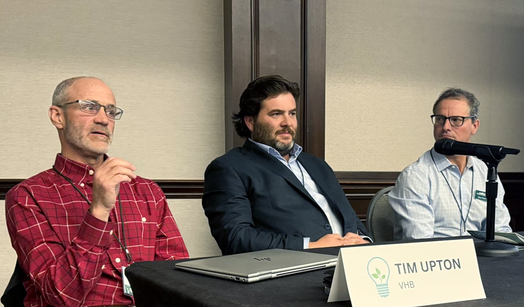 A panel of three individuals seated at a table with nameplates, speaking at an indoor event. 