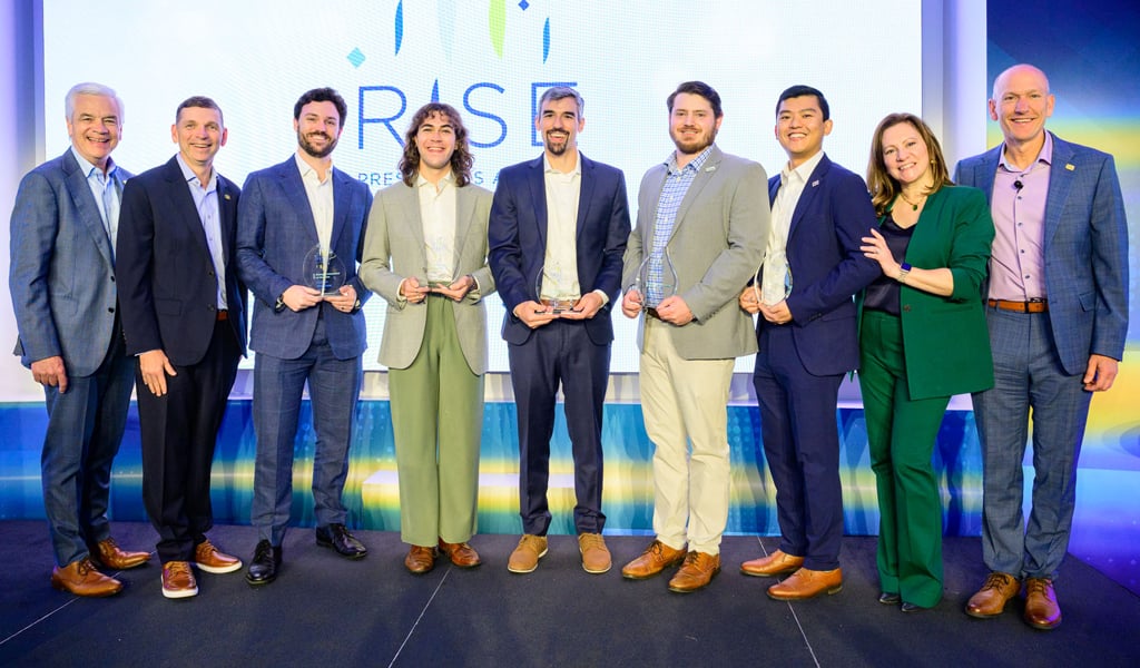 : Chair Mike Carragher, Chief Operating Officer Tom Jackmin, Darren Walshaw, Matt Burmeister, Eric Berger, Oliver Burke, Zachary Tiang, Chief People Officer Keri Kocur, and President Bill Ashworth stand on stage. The five award recipients in the center hold glass trophies.