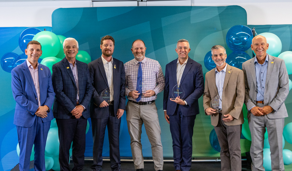 Seven people in business attire standing against teal and blue balloon backdrop, four holding glass awards.
