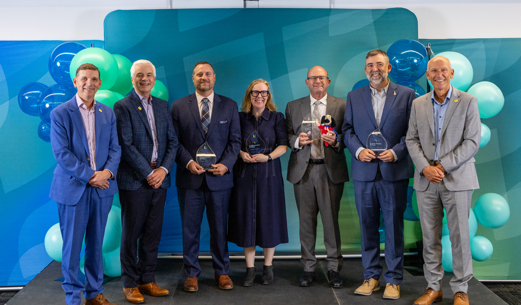 Seven people in business attire standing against teal and blue balloon backdrop, four holding glass awards.