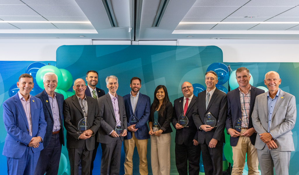 Eleven people in business attire standing against teal and blue balloon backdrop, seven holding glass awards.