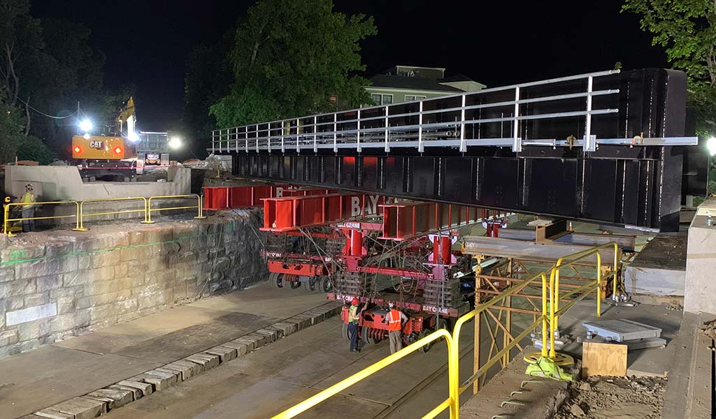 Ground level view of bridge replacement operation taking place at night. Workers, construction vehicles, heavy equipment and safety barricades are part of the scene.