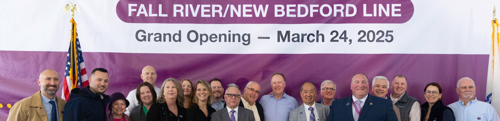 A large group of people standing between a purple banner announcing South Coast Rail service launch.
