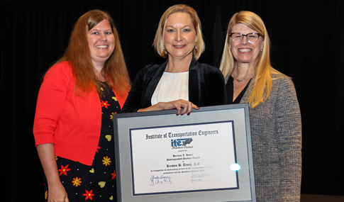 Kirsten Tynch stands in between two individuals smiling and holding her award plaque.