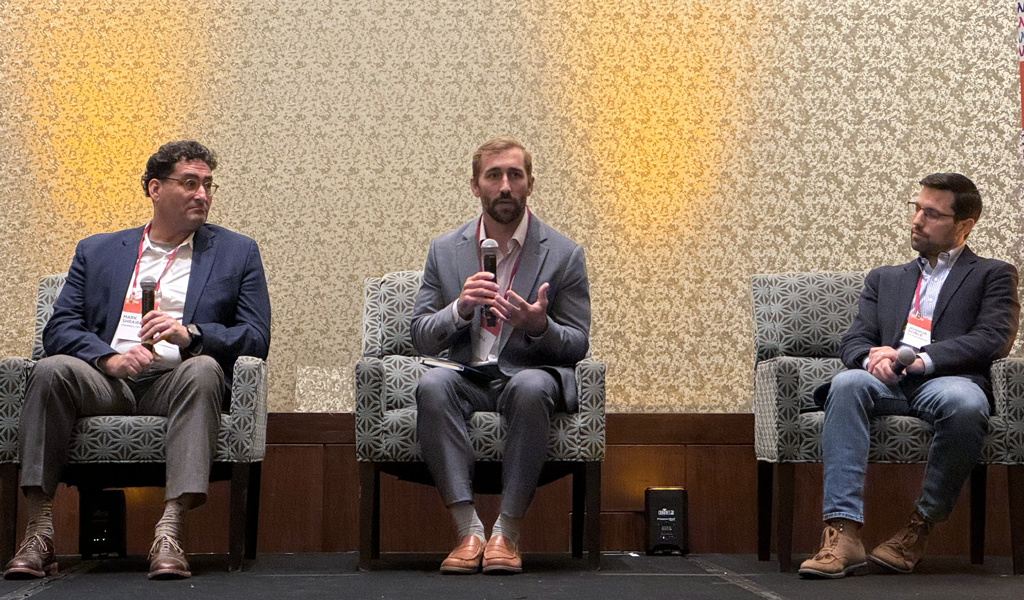 Three panelists, sitting on stage in a conference room, engage with the audience. The central panelist holds a microphone.