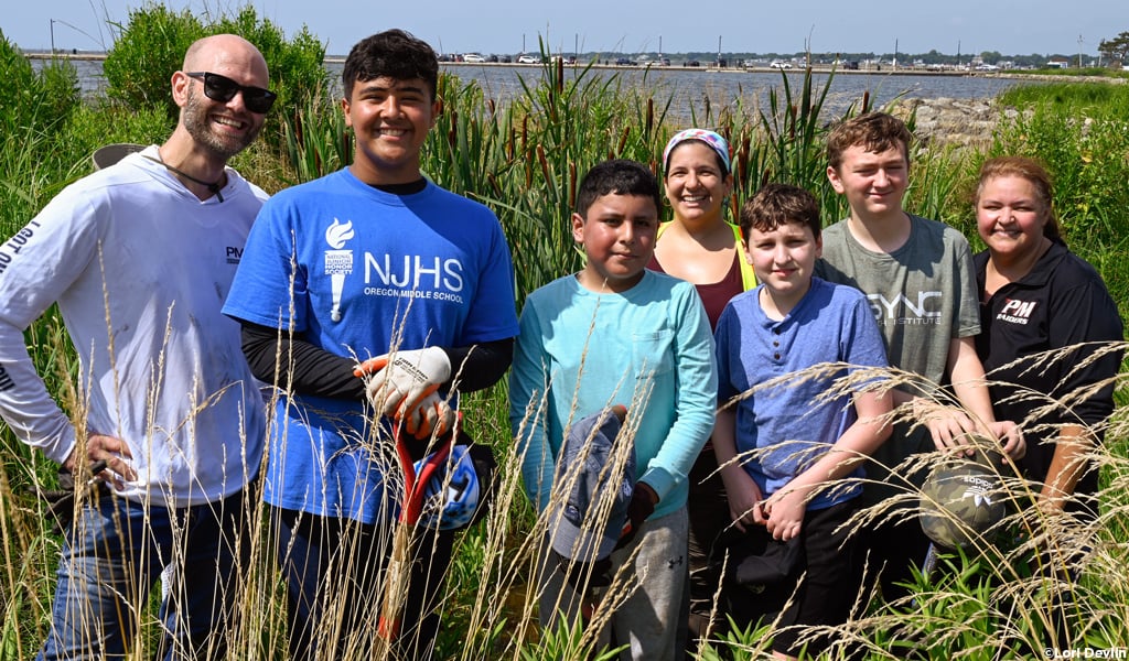 Six students and community volunteers stand in tall grass under a clear blue sky.
