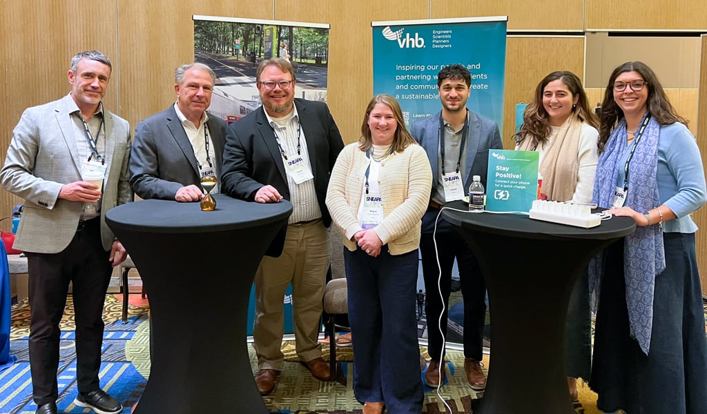 A group of seven people stands together indoors, smiling at a conference booth. The booth has a banner displaying the VHB company logo and information.  
