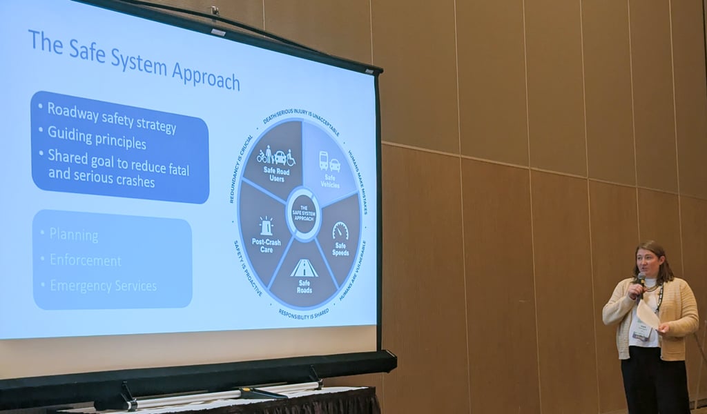 A person is giving a presentation in a conference room. The slide on display is titled "The Safe System Approach" and outlines a roadway safety strategy.