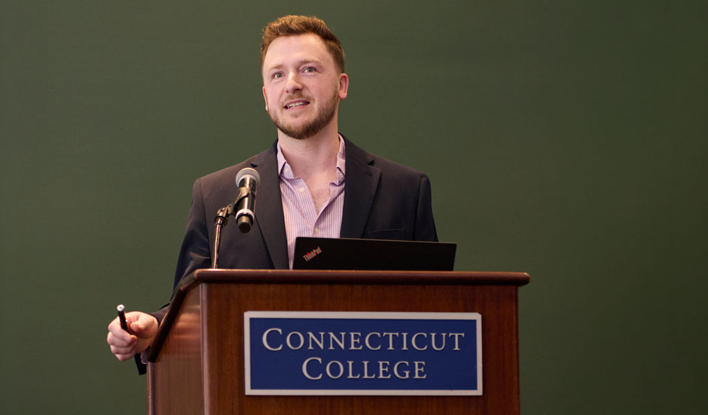 A person stands at a podium with a sign reading "Connecticut College," speaking into a microphone and holding a presentation clicker.
