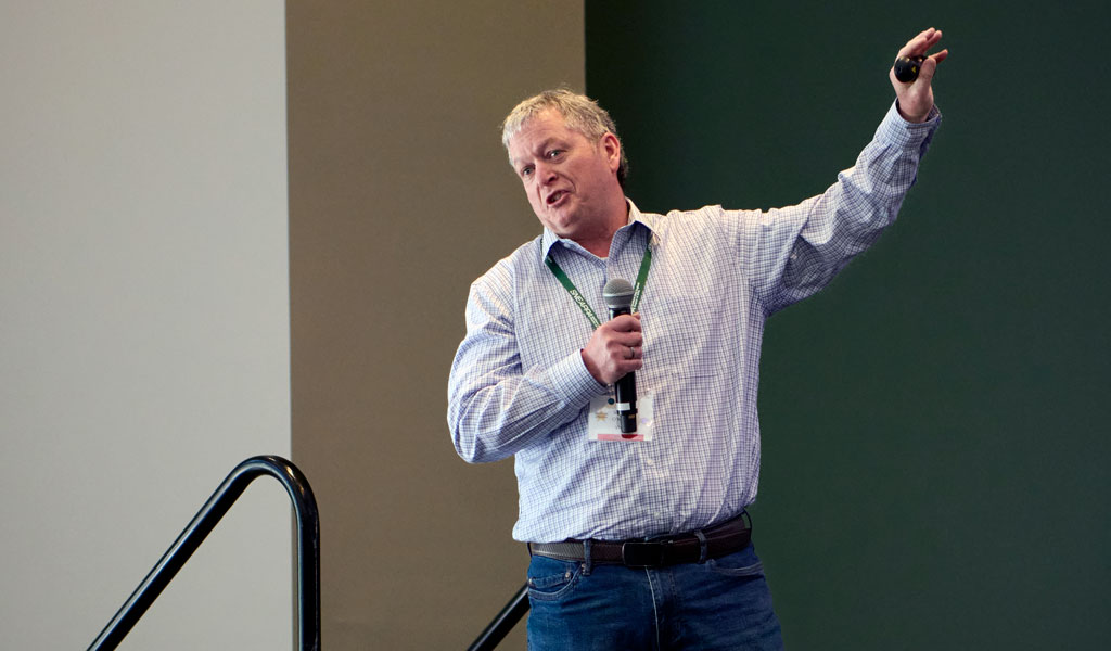 A person stands on a stage holding a microphone and pointing upwards. They are dressed in a checkered shirt and jeans.