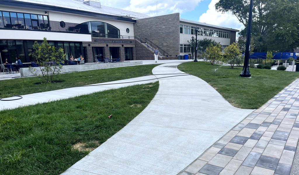 A paved pathway curves through a grassy area. There are small trees along the path and a modern building with large windows.