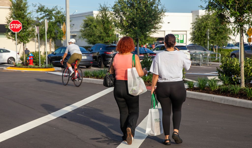 Two women with Sprouts bags walking in a crosswalk and a cyclist biking through the parking lot.
