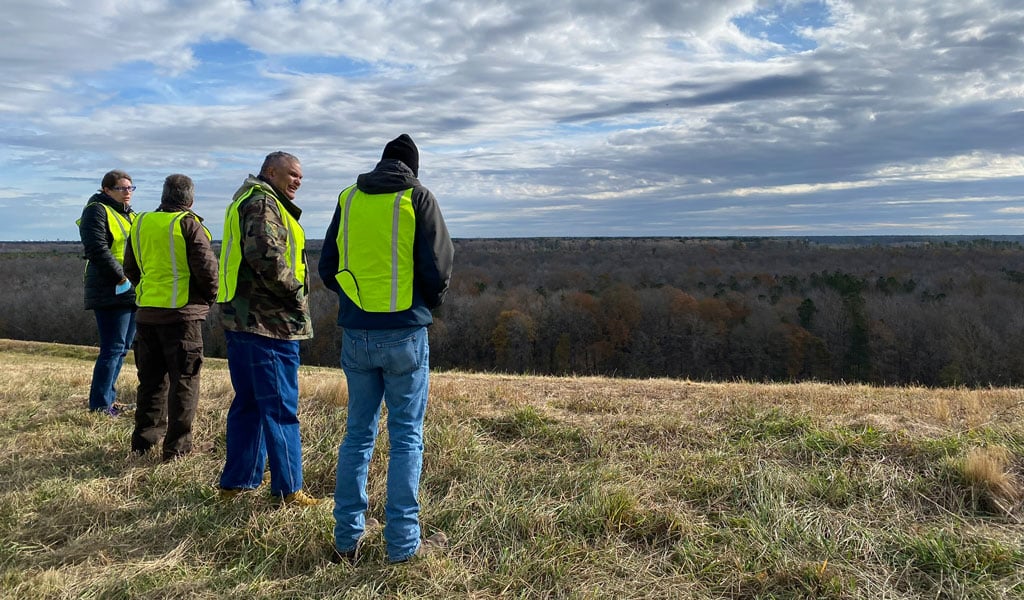 Tour participants in safety vests standing on a vegetated landfill cap, looking across the closed landfill area and surrounding forest.
