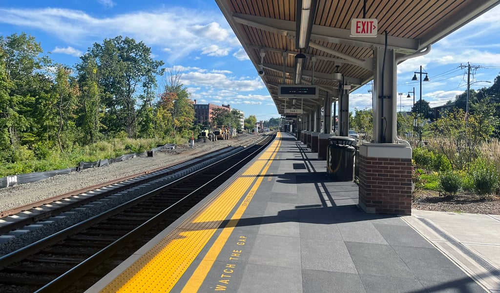 A train station platform with tracks on the left side. The platform has a yellow tactile strip along the edge, a covered waiting area with a wooden ceiling, and signage.