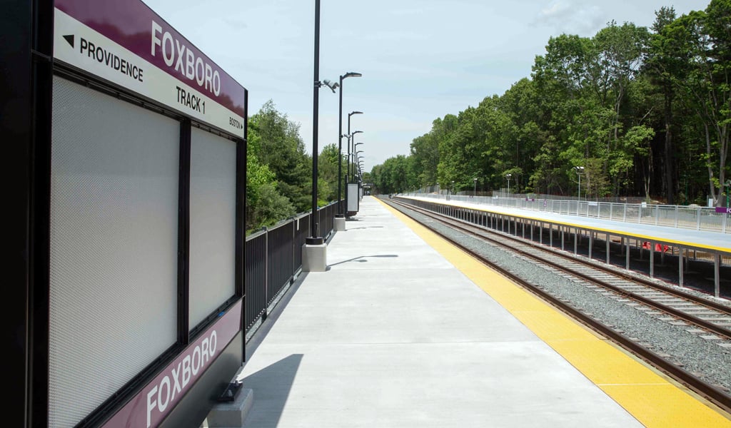 A train platform at Foxboro station is shown. The platform is empty with a sign indicating "PROVIDENCE" and "TRACK 1." Trees line the area alongside the tracks under a clear sky.