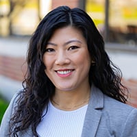 A person with long curly hair, wearing a light gray blazer and a white shirt, is positioned against a black background.