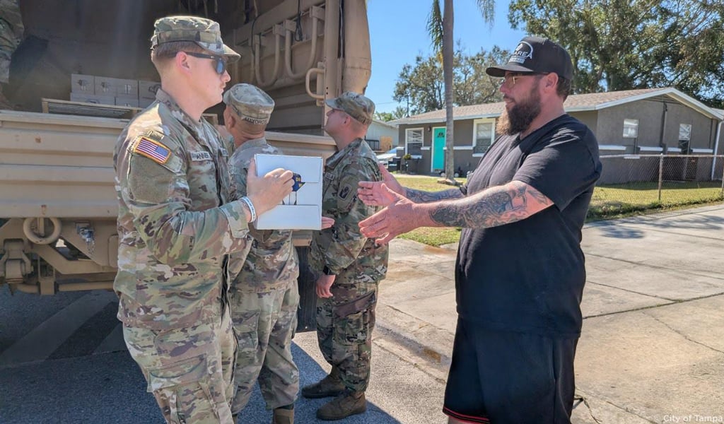 Military personnel unload storm recovery supplies to a man wearing a black hat.