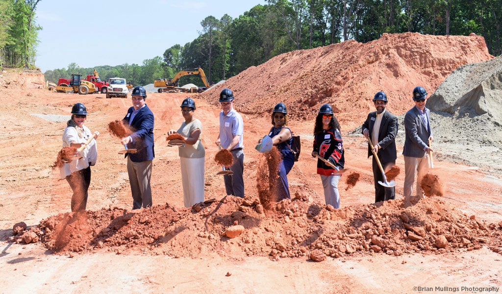 People wearing hard hats standing in line on construction site, holding shovels, tossing soil at groundbreaking.