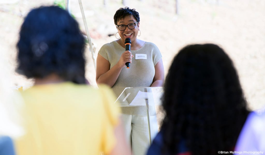 Woman smiling, standing at podium, holding microphone speaking to audience.