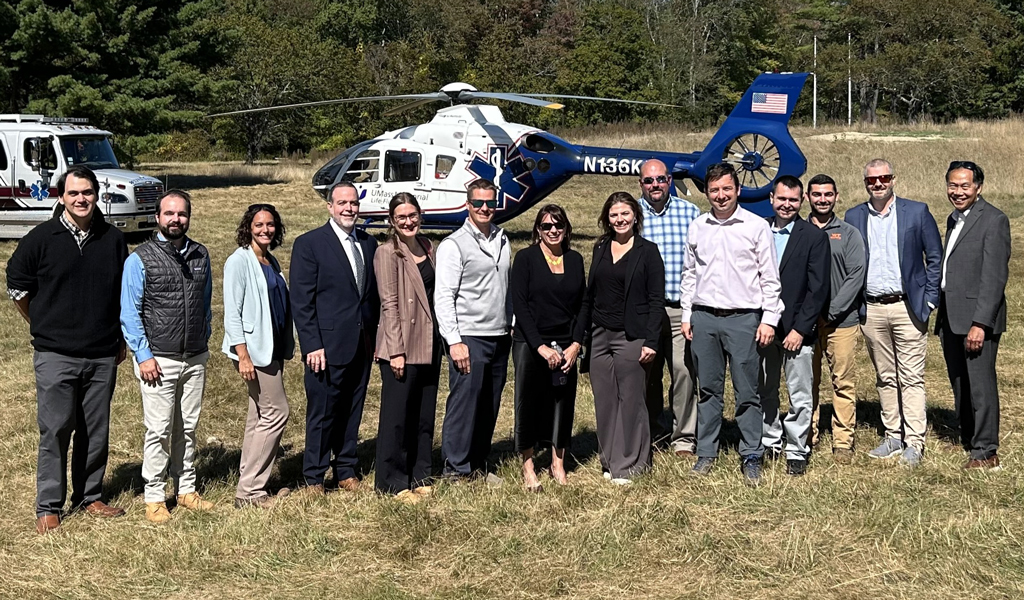 Group of professionals standing in a field with a helicopter and emergency vehicles in the background.