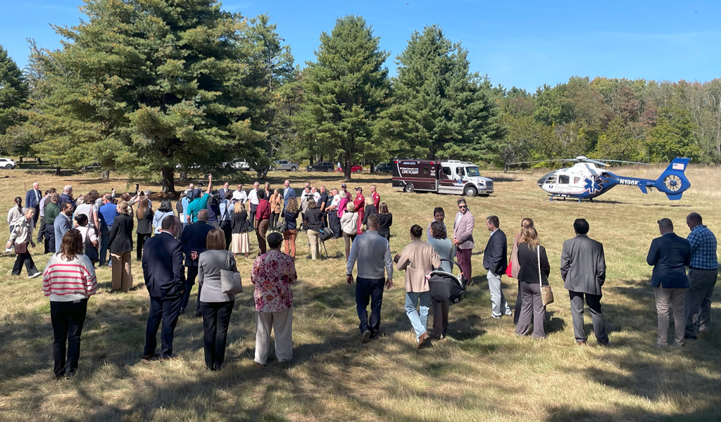 Group of people gathered outdoors for an event with emergency vehicles and a helicopter nearby on a sunny day with trees in the background.